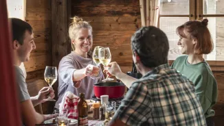 Deux femmes et deux hommes sont assis ensemble à une table dans un chalet en bois et dégustent une fondue au fromage. Ils trinquent avec un verre de vin blanc.